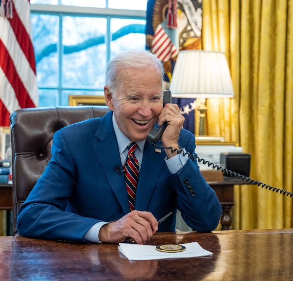 President Biden smiles while speaking on a telephone in the Oval Office.
